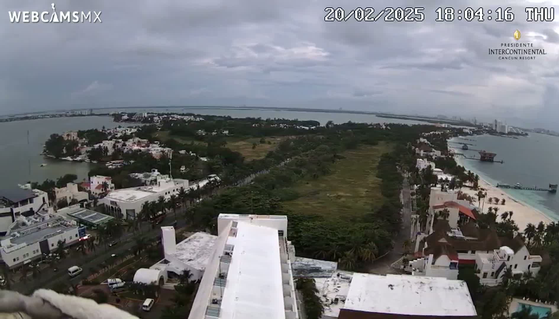 An aerial view shows a coastal city under an overcast sky, featuring a beach, buildings, and a large body of water.