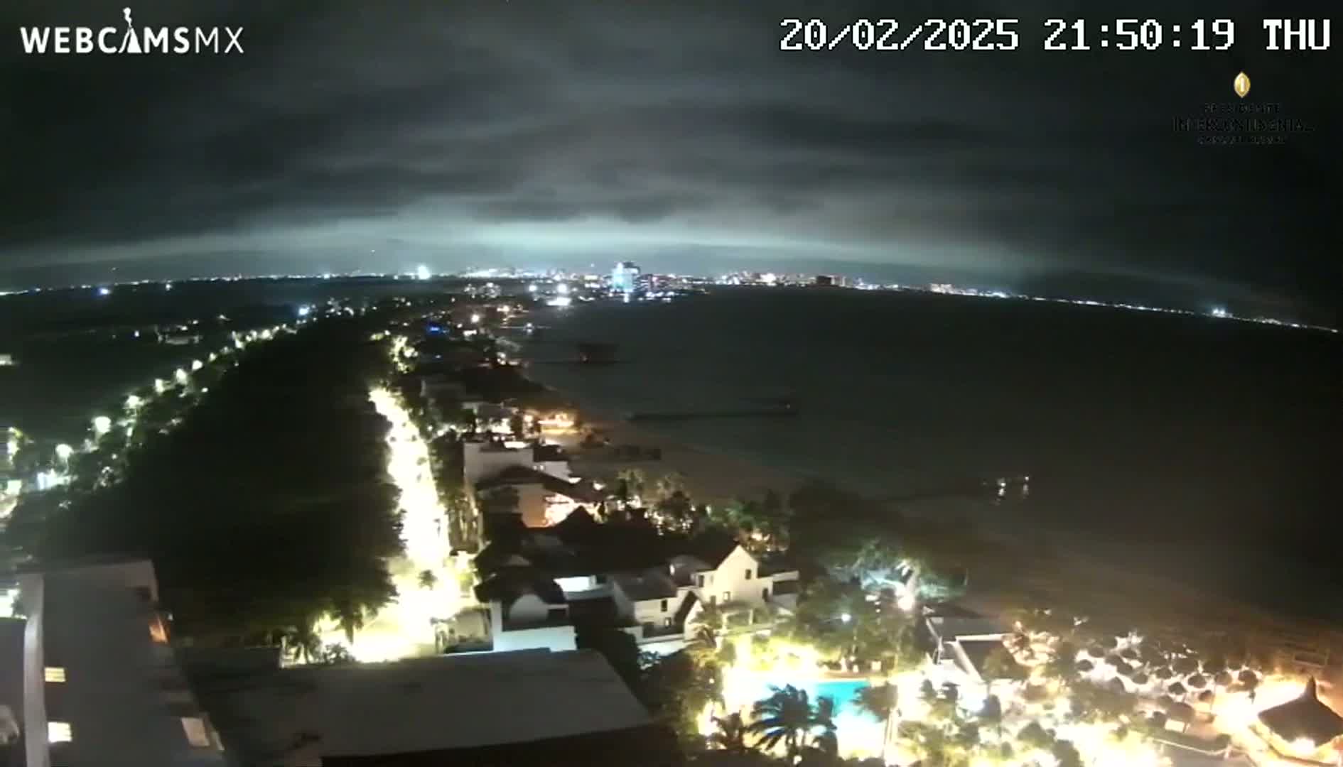 A nighttime coastal scene shows a city skyline across the water, with a beach, buildings, and palm trees illuminated along the shore under a cloudy sky.