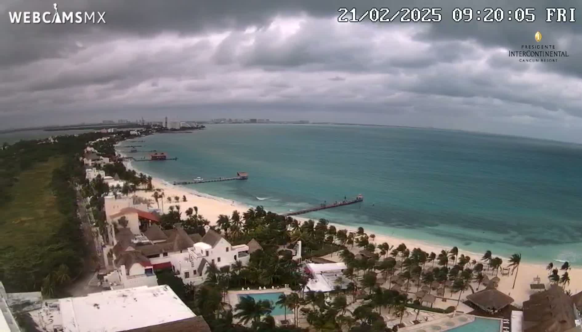 An aerial view shows a tropical beach with overcast skies, a long pier extending into turquoise water, and beachfront buildings under a cloudy sky.