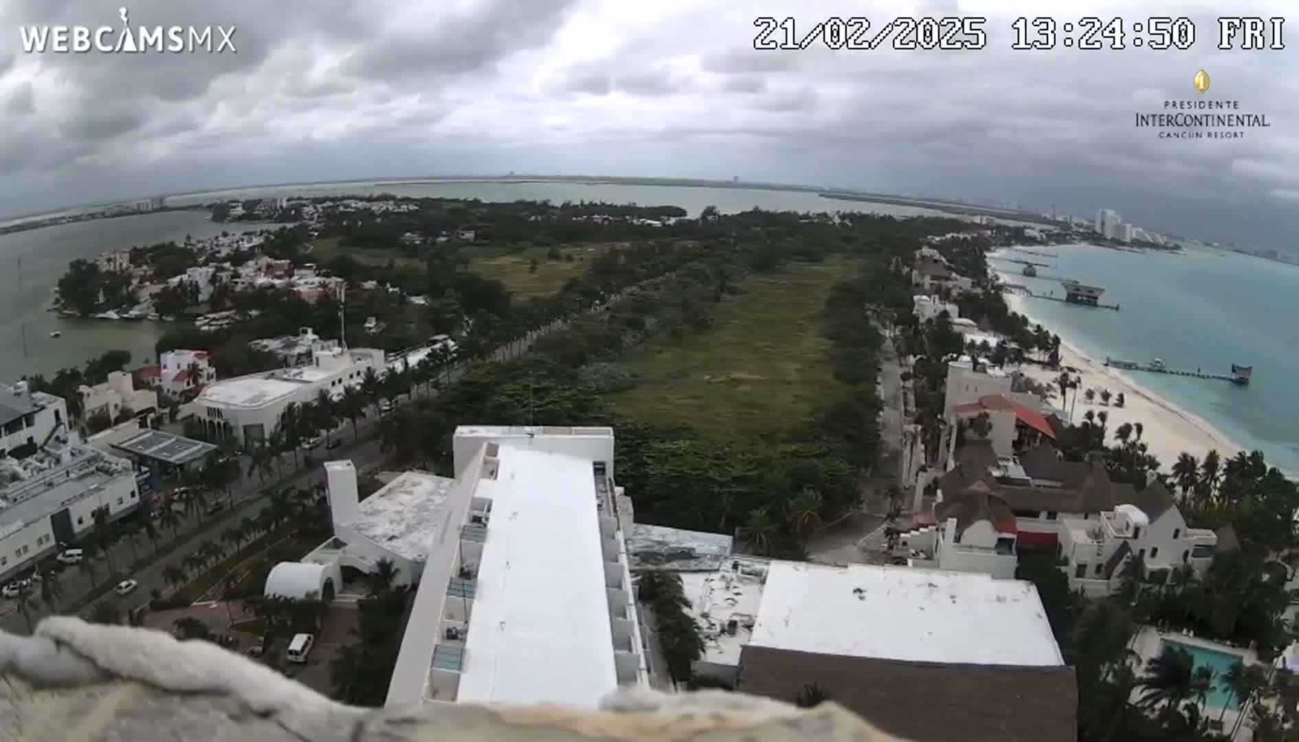 An elevated view shows a coastal city under an overcast sky, featuring buildings, roads, a beach, and a body of water.