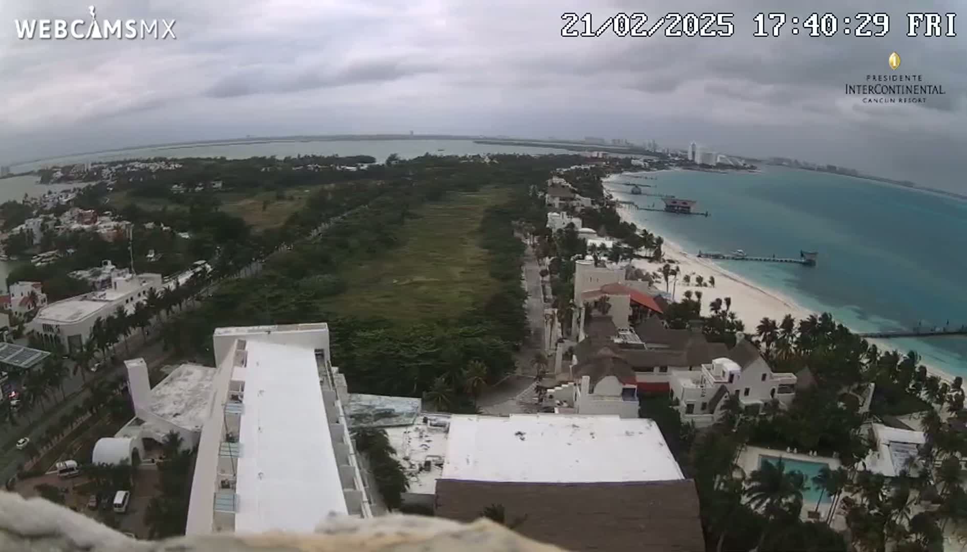 An aerial view shows a beachside resort town on an overcast day, with a stretch of sandy beach, turquoise water, and low-lying buildings.