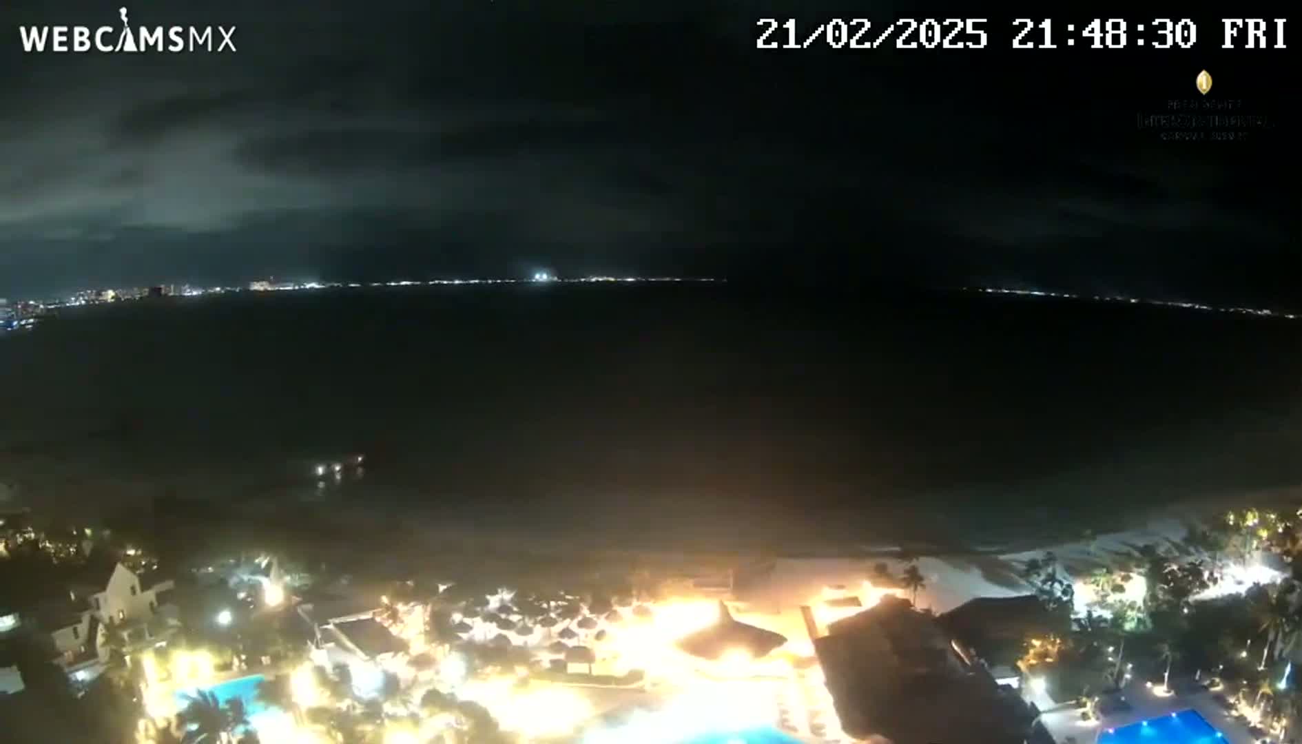 A nighttime view of a coastline shows a dark ocean, a brightly lit resort, and a distant city skyline under a mostly cloudy sky.