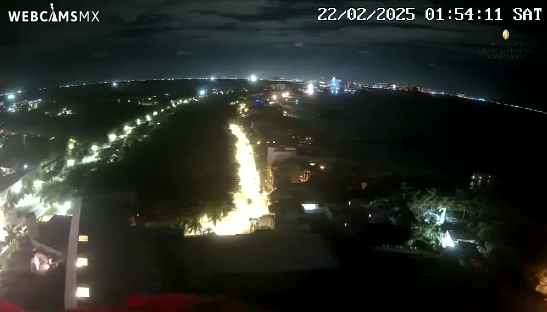 A nighttime aerial view shows a brightly lit road winding through a dark, tree-lined area toward a distant, illuminated cityscape under a mostly clear sky.