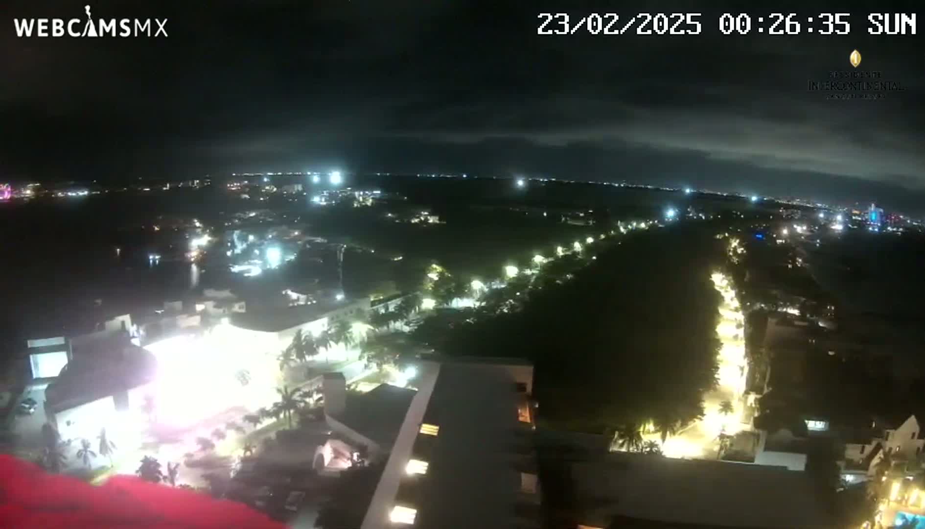 A nighttime, high-angle view shows a city illuminated with streetlights, a body of water in the distance, and a portion of a building in the foreground under a partly cloudy sky.