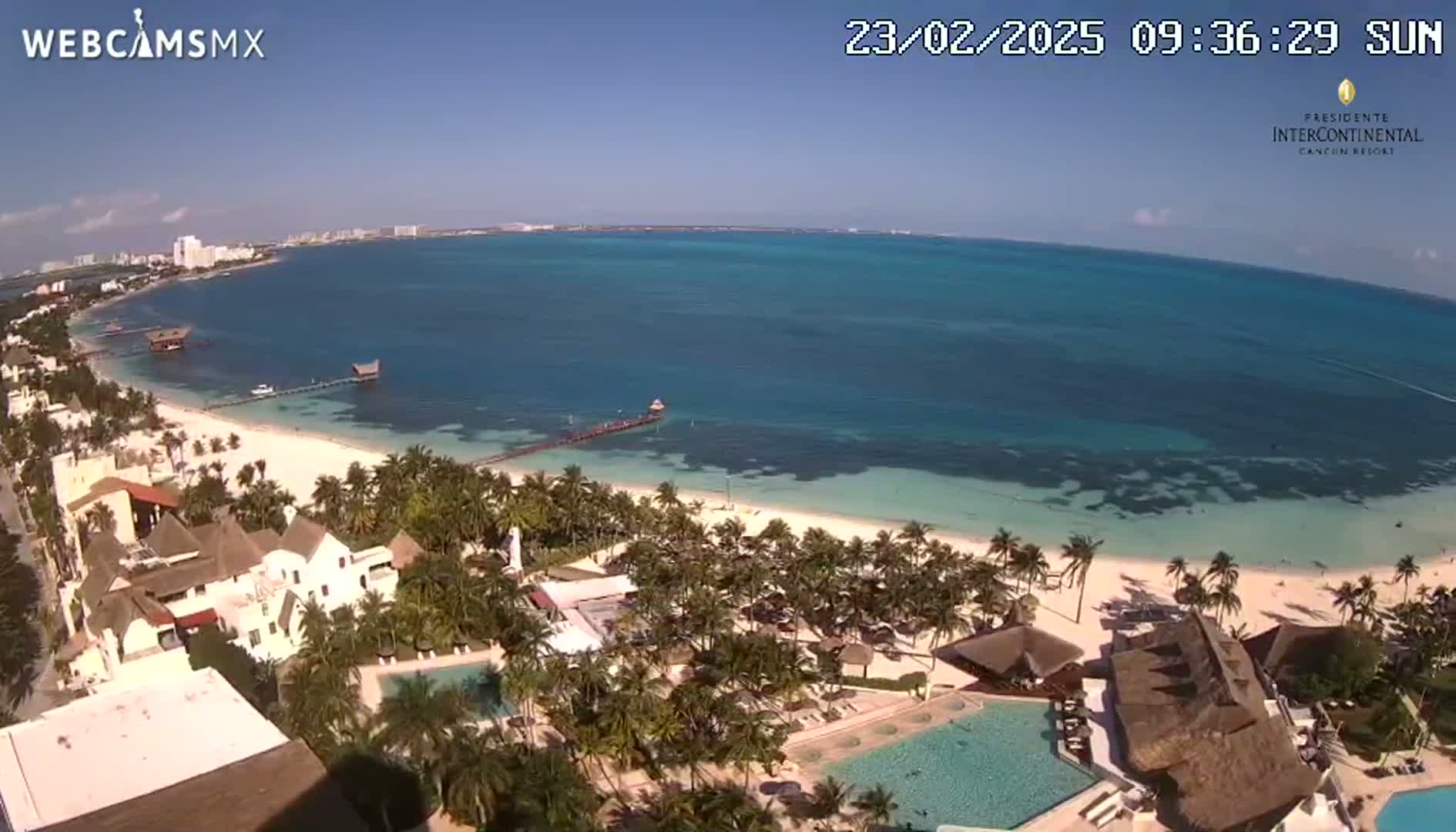 An aerial view shows a resort on a white sand beach next to a calm, turquoise ocean under a clear blue sky.