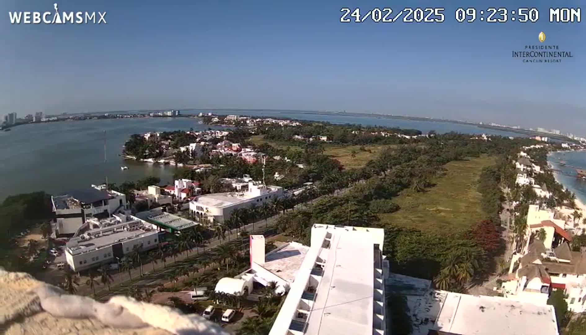 An aerial view shows a coastal town on a sunny day, with a body of water and a peninsula in the background.