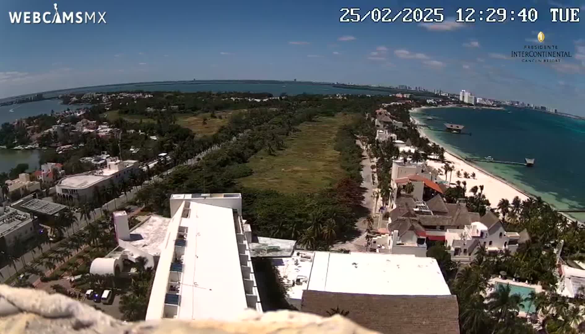 An aerial view shows a sunny beach, ocean, and coastal buildings, with a large expanse of undeveloped land separating the beach from the buildings further inland.