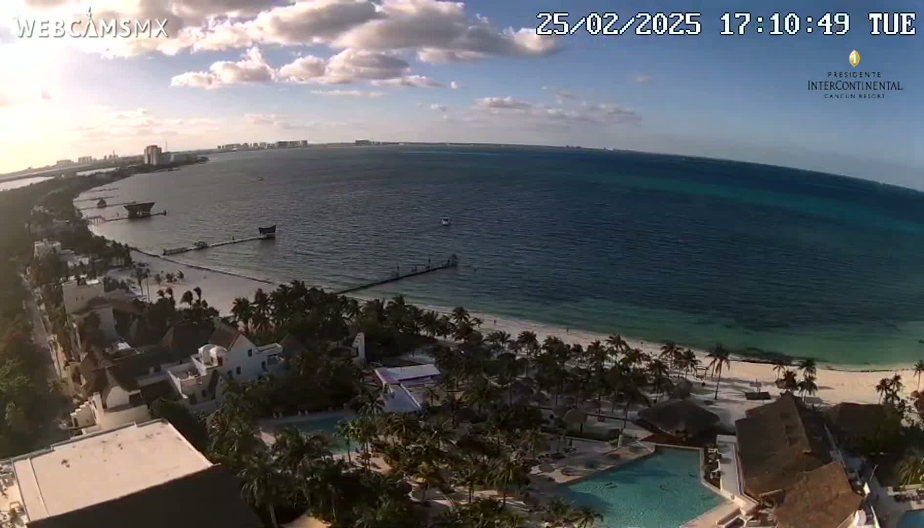 An aerial view of a beach resort on a partly cloudy day shows a calm ocean, white sand beach, palm trees, and resort buildings with swimming pools.