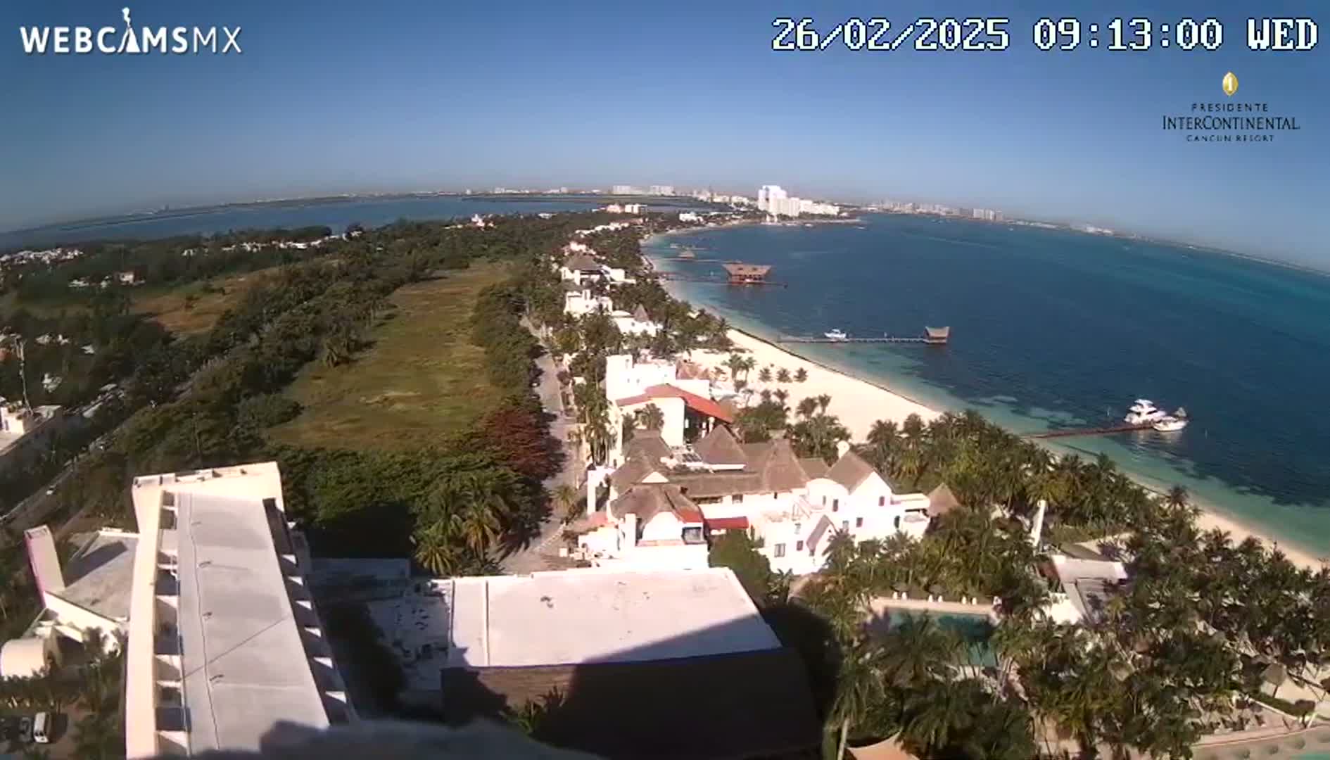 An aerial view shows a tropical beach with white sand, turquoise water, palm trees, and low-rise buildings on a sunny day.