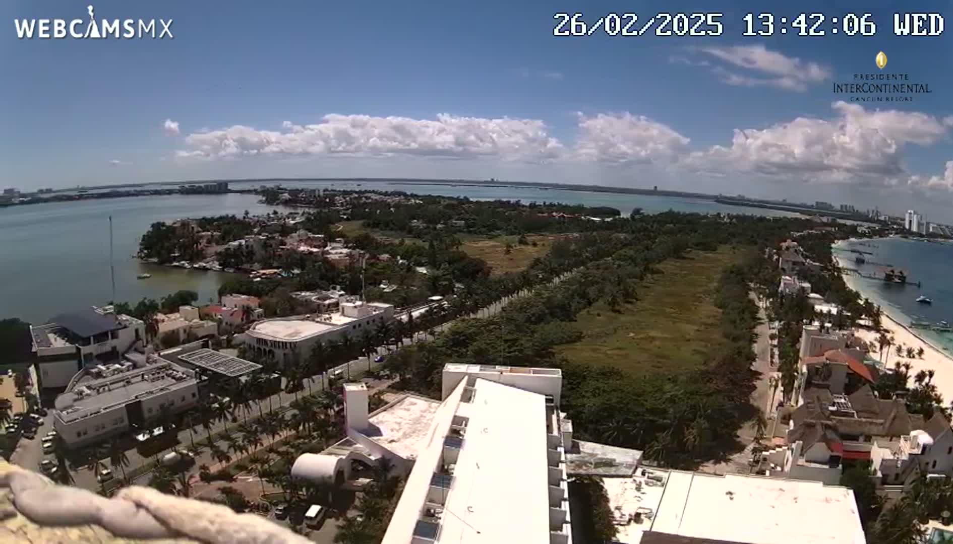 An aerial view shows a coastal city on a partly cloudy day, with a body of water and a beach visible in the distance.