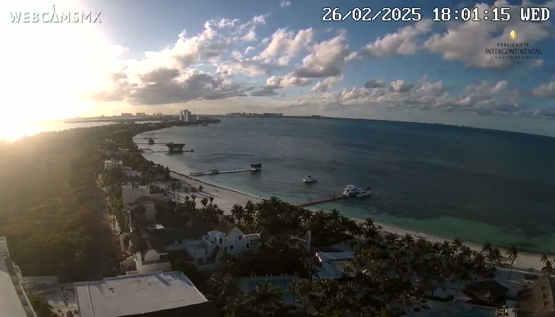 A mostly sunny, partly cloudy sunset view of a tropical beach with calm ocean waters, palm trees, and buildings.