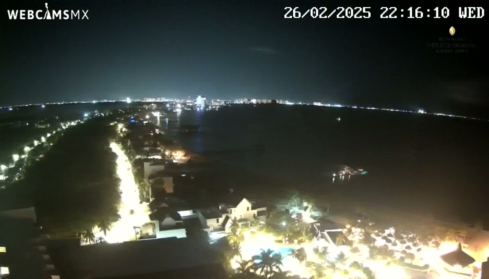 A nighttime, high-angle view shows a coastal city with illuminated buildings, roads, and palm trees, overlooking a dark body of water with a distant city skyline on the horizon.