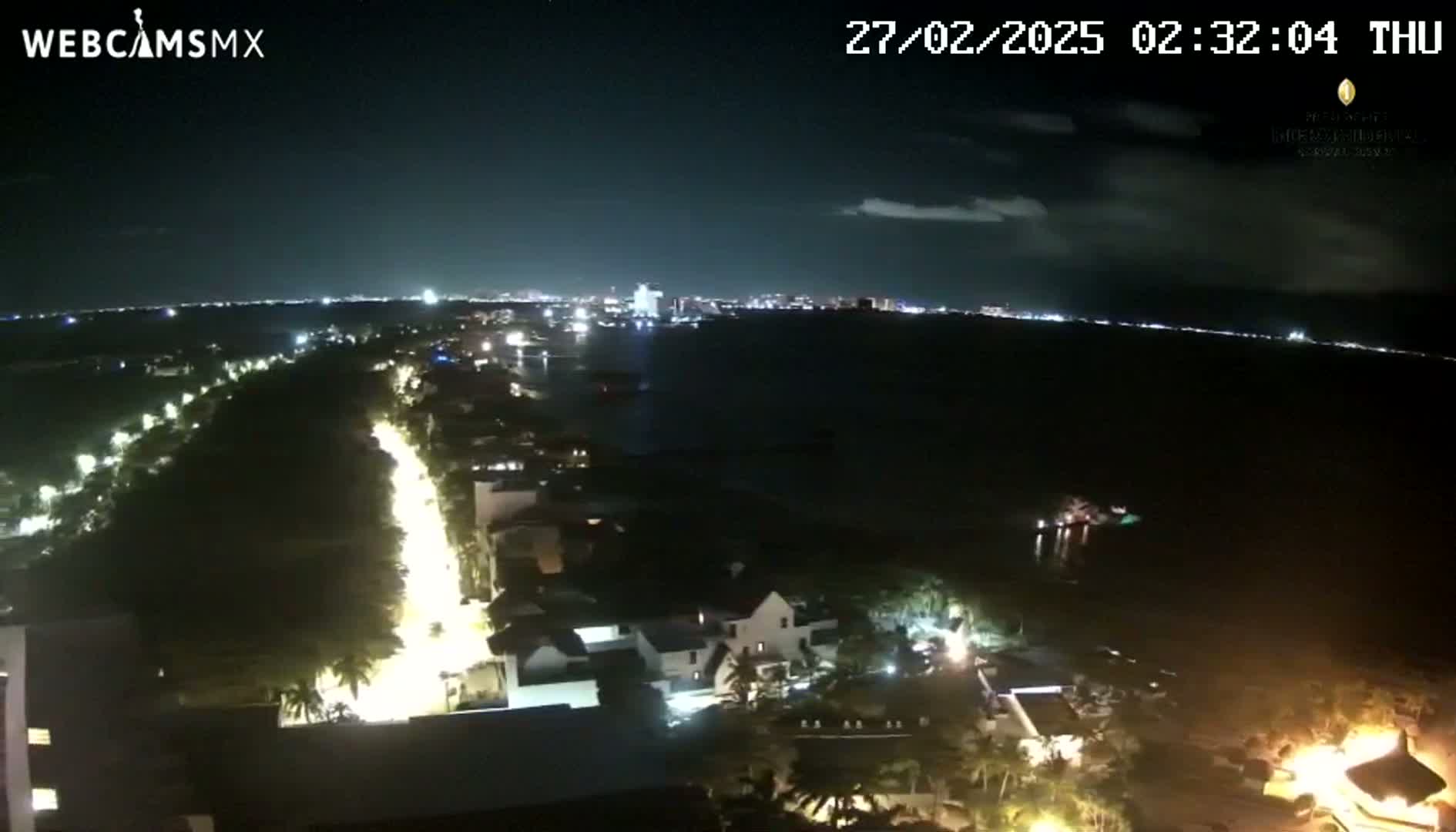 A nighttime, high-angle view shows a coastal town with illuminated streets and buildings next to a dark body of water, with a city skyline visible in the distance.
