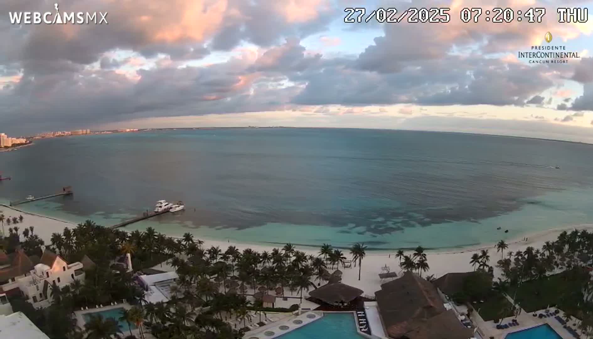 An aerial view shows a calm ocean with a white sandy beach lined with palm trees and resort buildings under a partly cloudy sky.