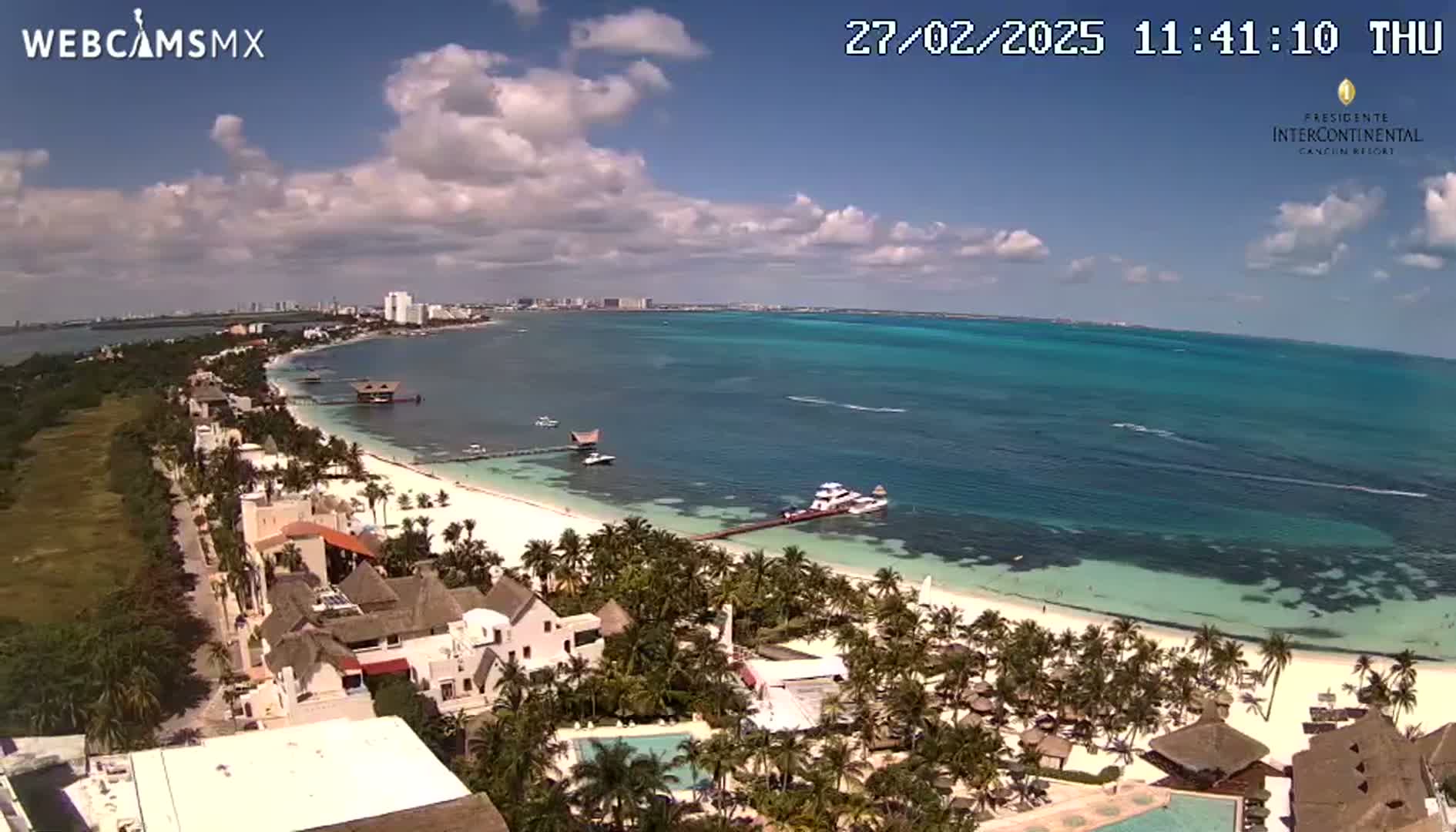 An aerial view shows a partly cloudy day over a tropical beach with turquoise water, palm trees, and resort buildings.
