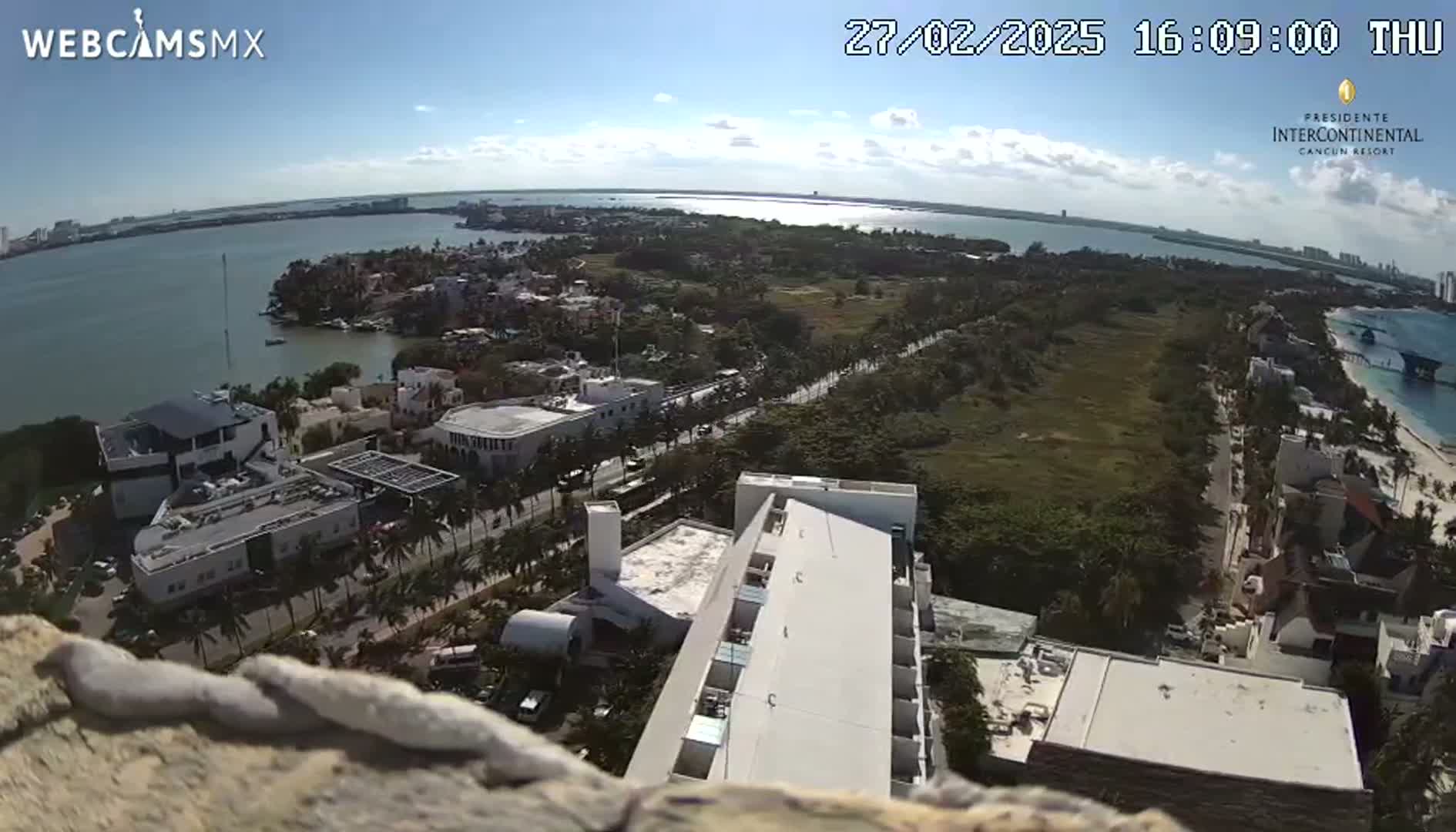 An elevated view of a coastal city on a sunny day shows buildings, roads, and a body of water with a peninsula jutting out.