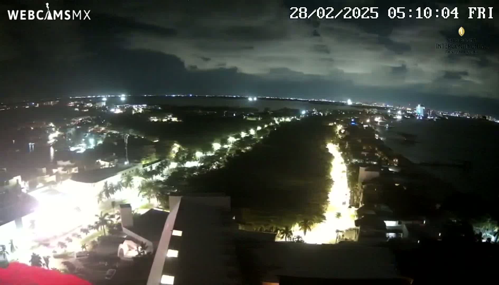 A nighttime, high-angle view shows a city illuminated with streetlights, a body of water in the distance under a cloudy sky.
