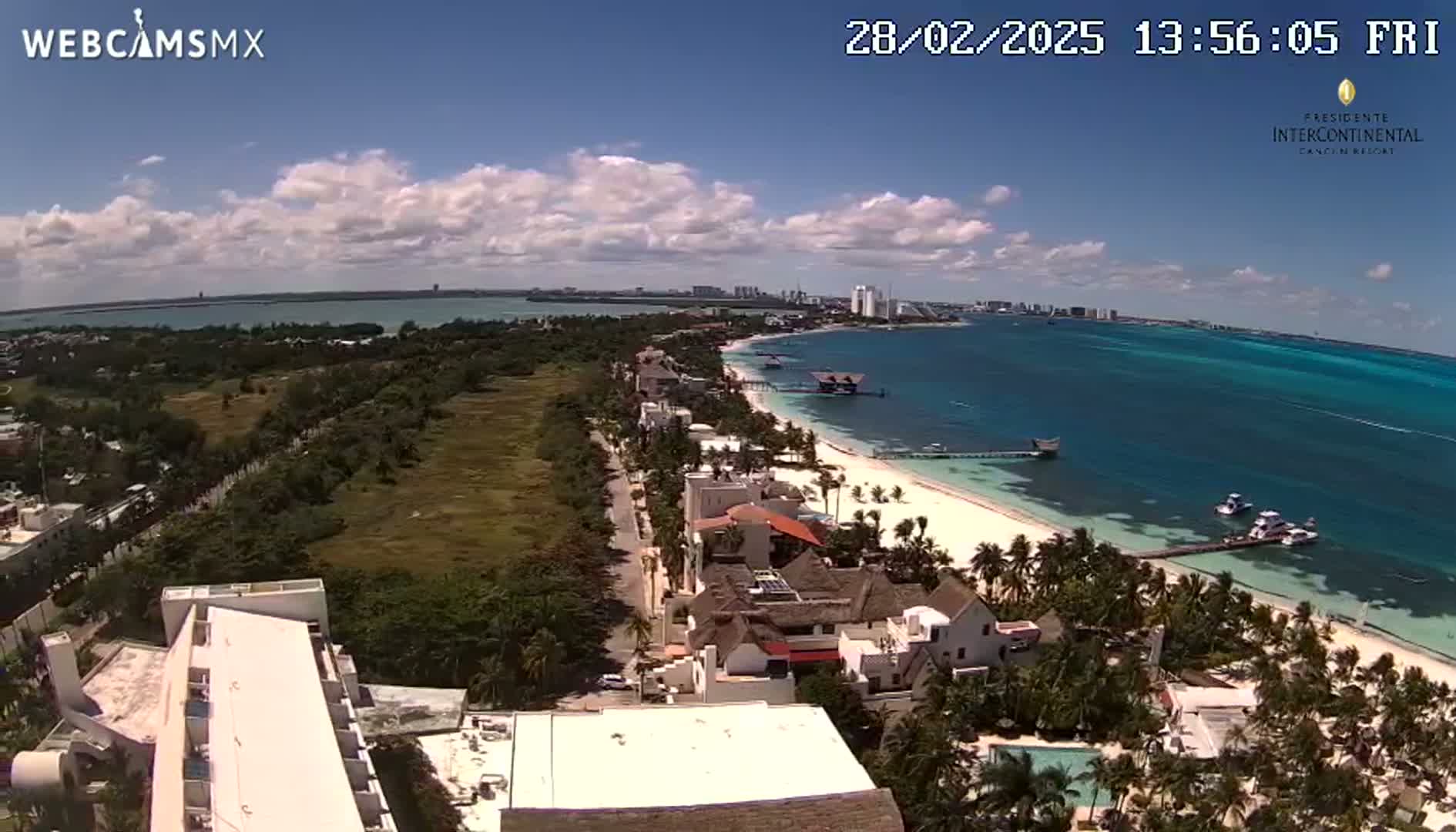 An aerial view shows a partly cloudy day over a tropical coastline with white sand beaches, turquoise water, and buildings along the shore.
