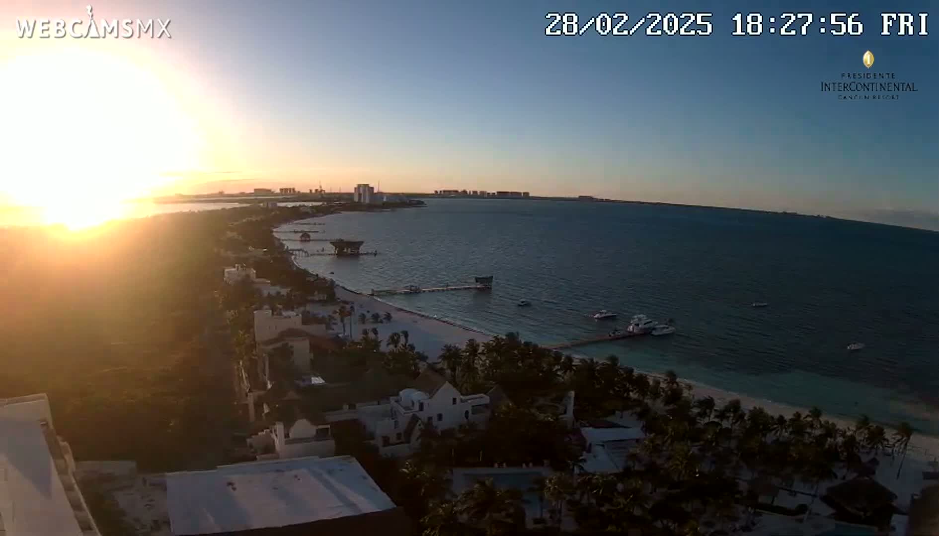 A coastal sunset scene shows a beach with palm trees and buildings, overlooking calm water with boats, under a clear sky.