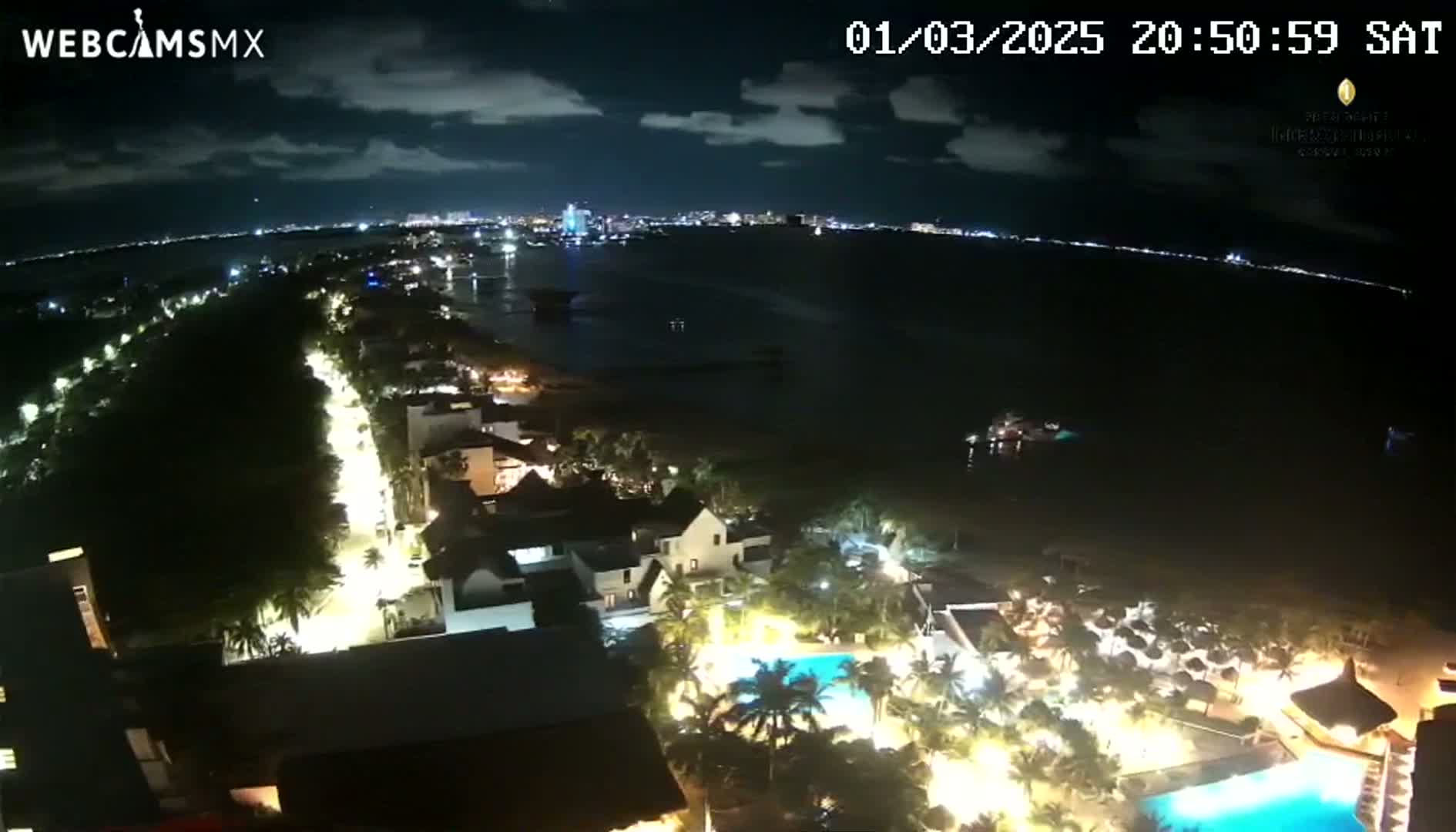 A nighttime aerial view of a coastal resort town shows a dark ocean, a brightly lit beachfront road, and numerous illuminated buildings and pools.