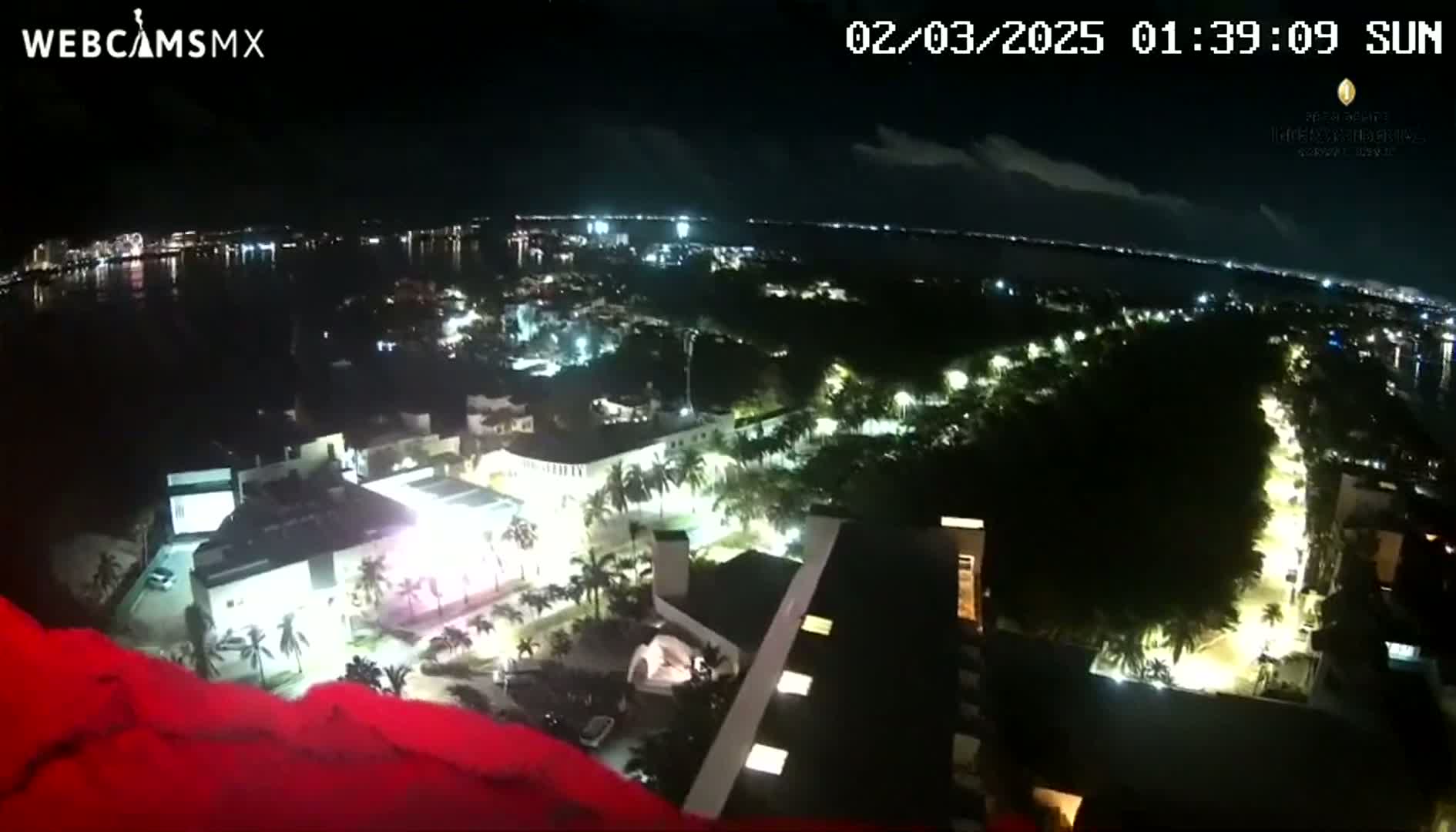 A nighttime aerial view of a coastal city illuminated with lights, under a partly cloudy sky, shows a body of water in the distance.