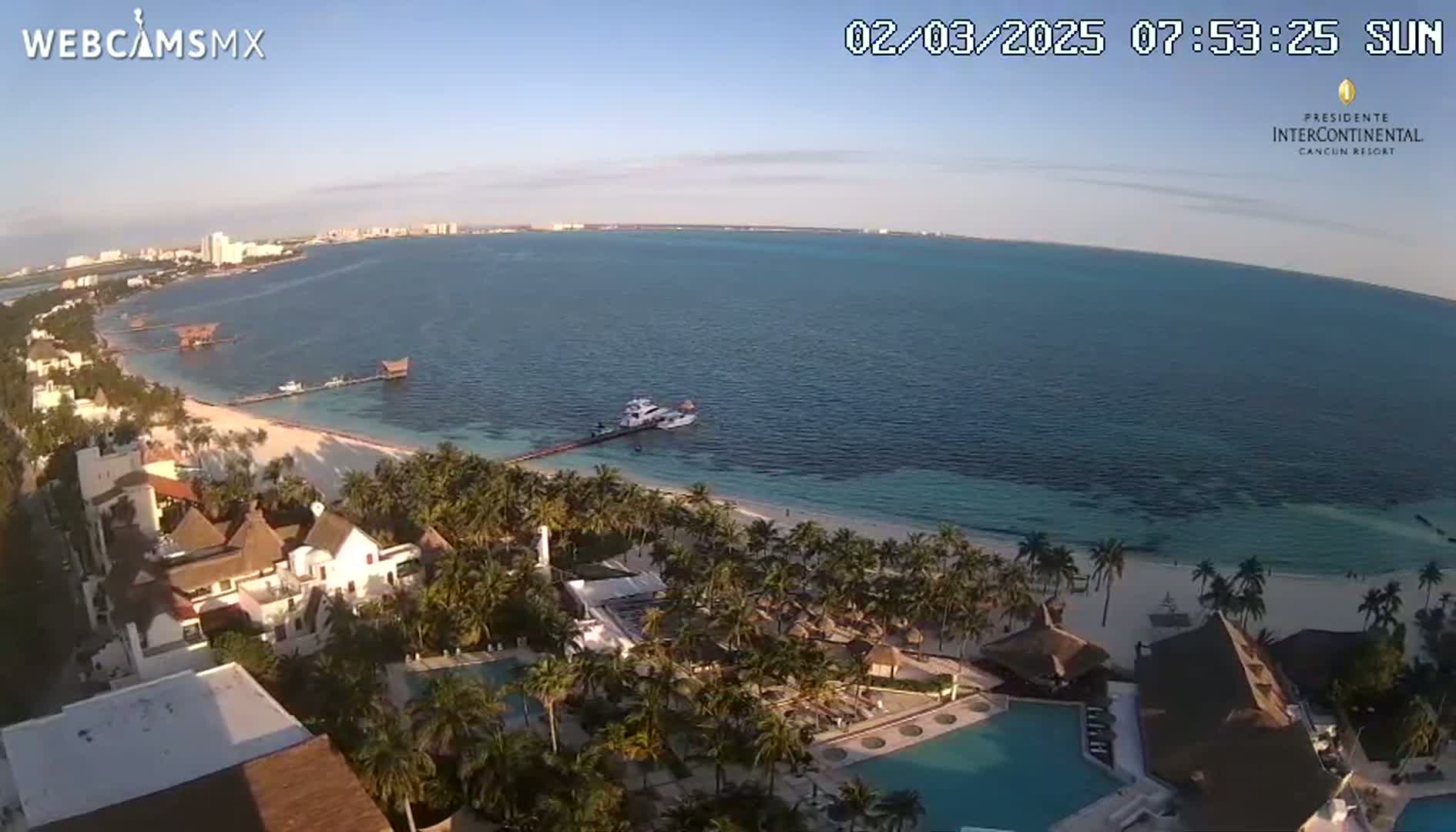 An aerial view shows a resort on a white sand beach next to a calm, turquoise ocean under a clear, sunny sky.