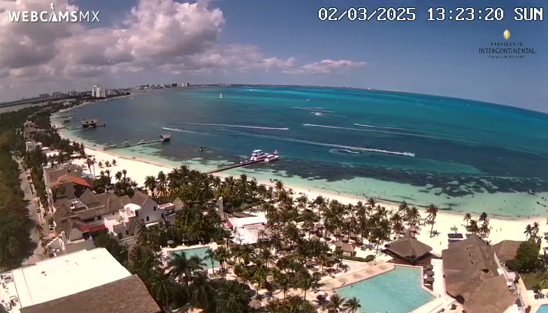 An aerial view shows a beach resort with pools, palm trees, and a white-sand beach bordering a turquoise ocean under partly cloudy skies.
