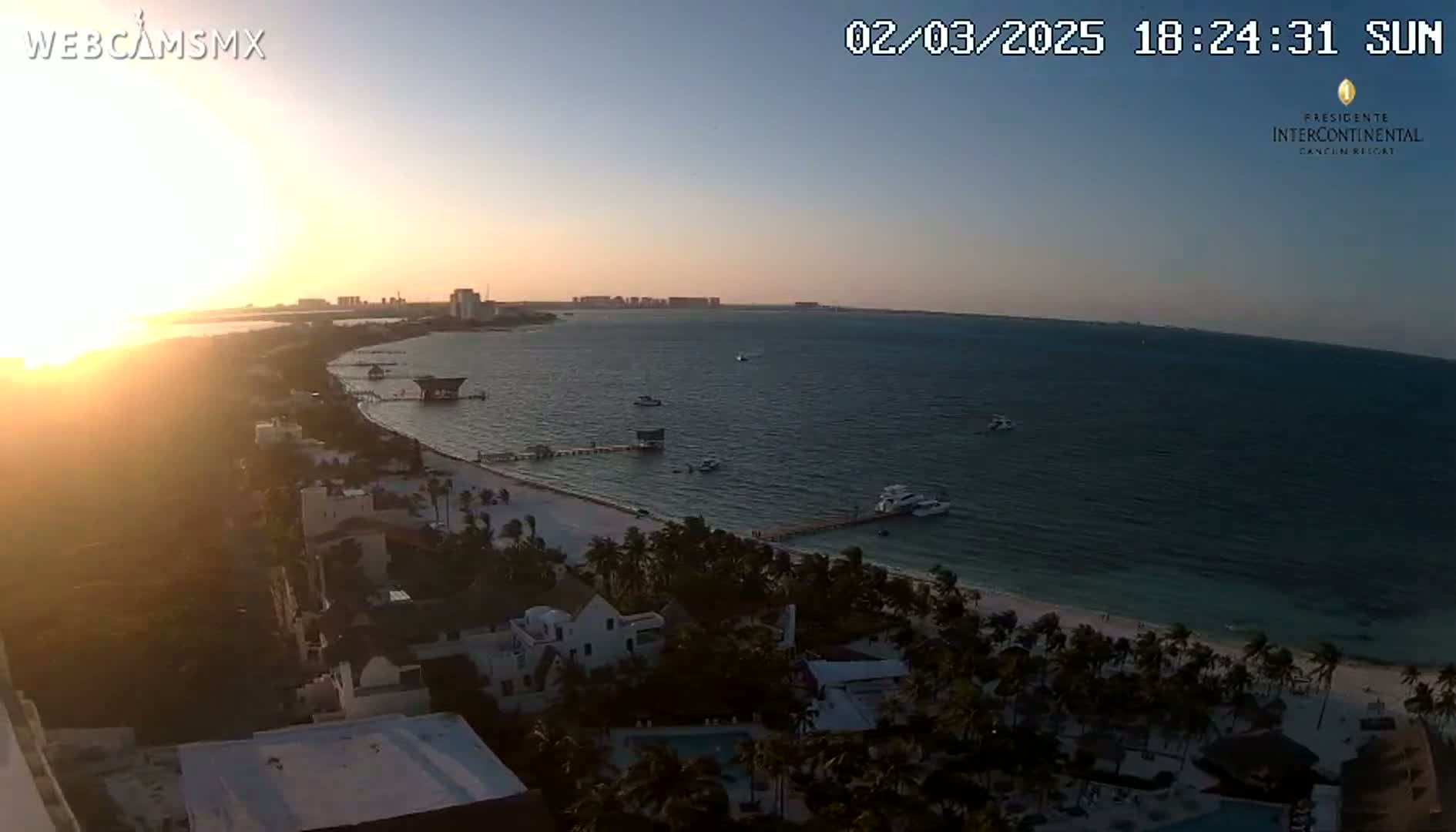 A sunset over a calm ocean, with a sandy beach and beachfront buildings visible, showing a clear sky.