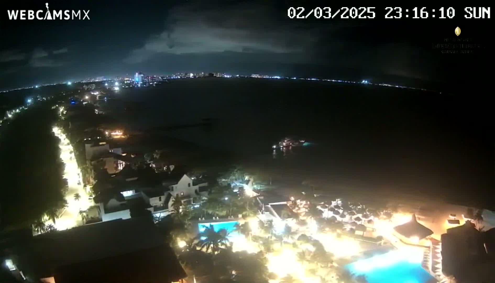 A nighttime aerial view shows a brightly lit resort area next to a dark ocean under a partly cloudy sky.