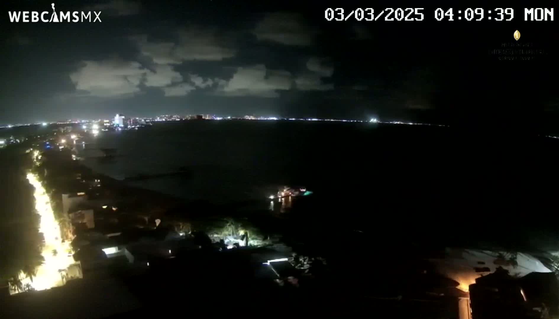 A nighttime coastal view shows a city skyline across a dark body of water, with a brightly lit street running along a shoreline under a partly cloudy sky.