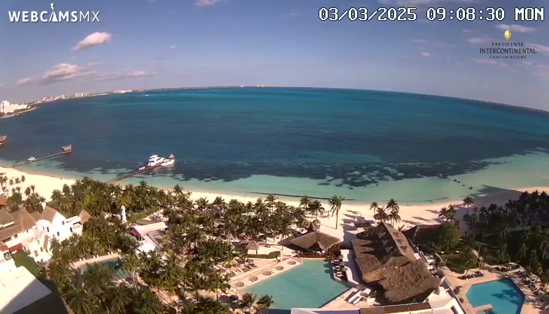 An aerial view shows a tropical resort with swimming pools, lush vegetation, and a pristine beach bordering a calm, turquoise ocean under a sunny sky.
