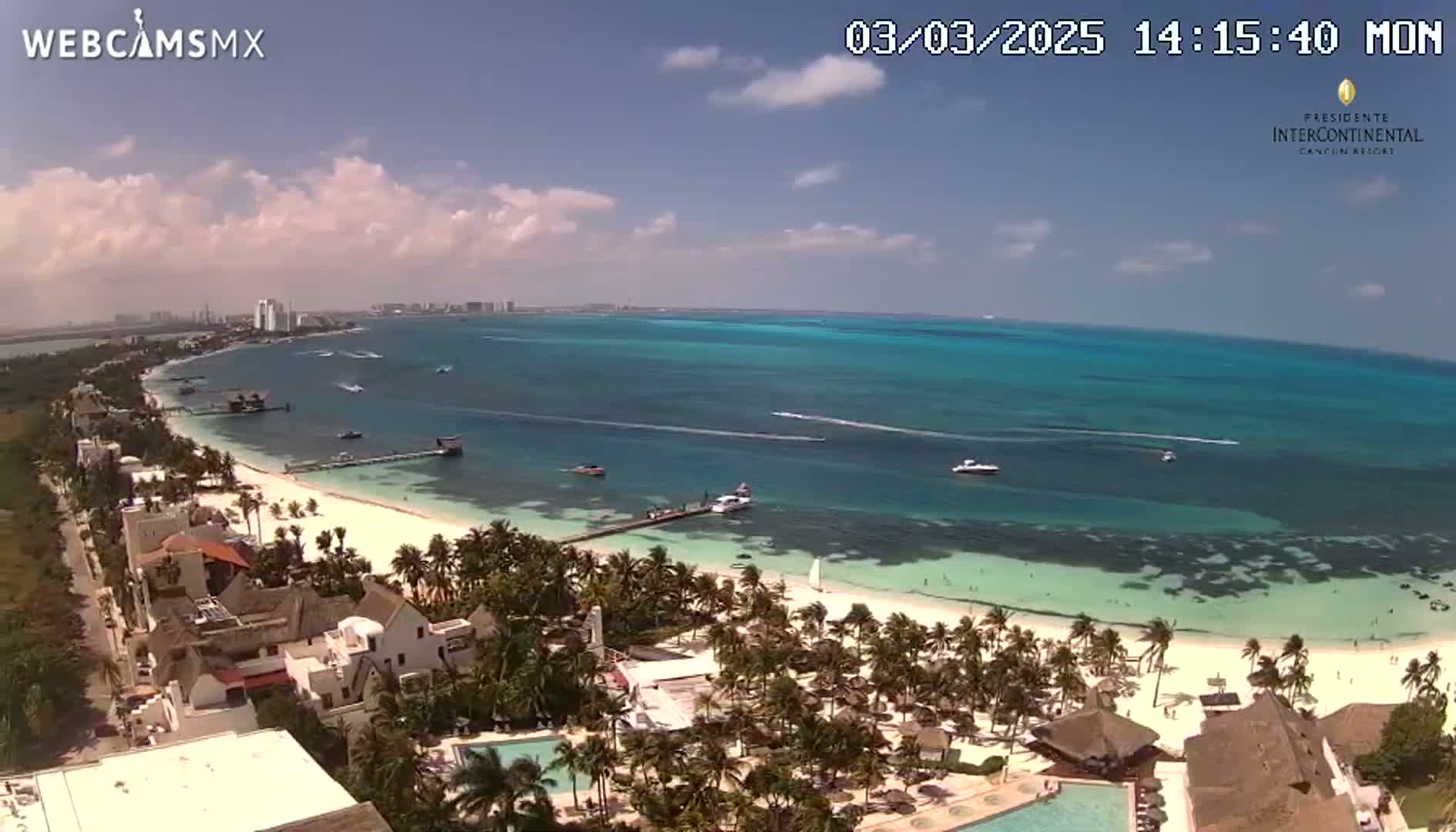 An aerial view shows a beach with turquoise water, palm trees, buildings, and a partly sunny sky.