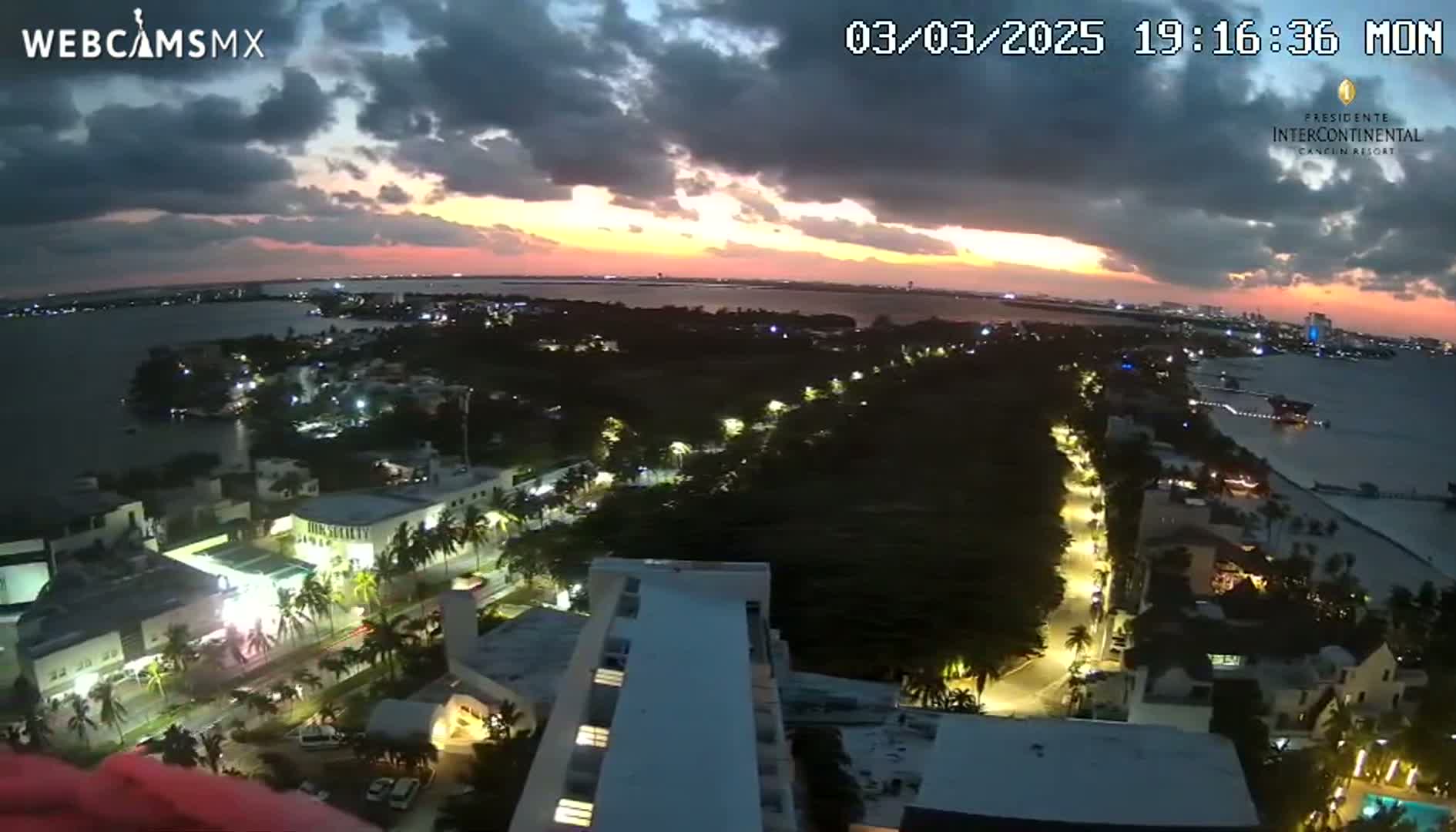 An aerial view shows a coastal city at dusk under a partly cloudy sky, with city lights illuminating streets and buildings next to a body of water.