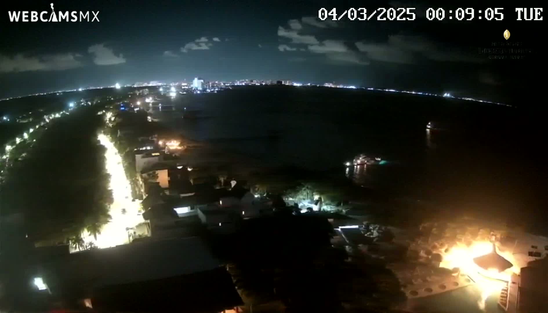 A nighttime aerial view shows a coastal town with a road illuminated by streetlights, a body of water with boats, and a fire burning near the waterfront under a partly cloudy sky.