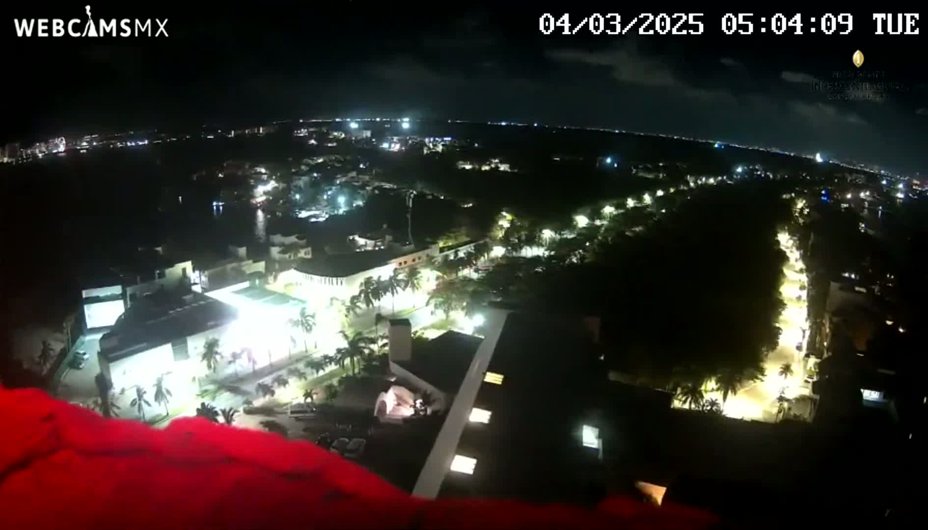 A nighttime aerial view of a city illuminated by streetlights and building lights, with a body of water visible in the distance under a clear sky.