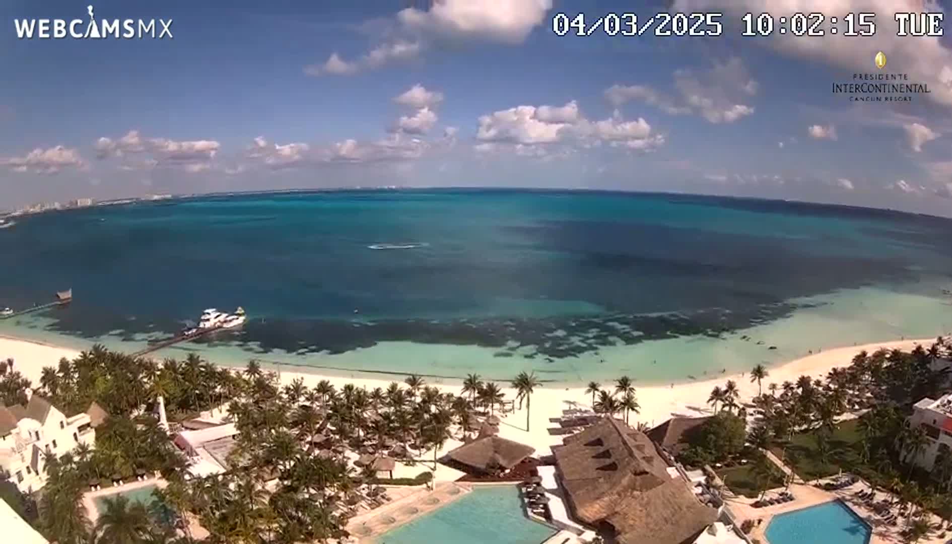 A tropical beach resort with a swimming pool overlooks a calm, turquoise ocean under a mostly sunny sky with a few scattered clouds.