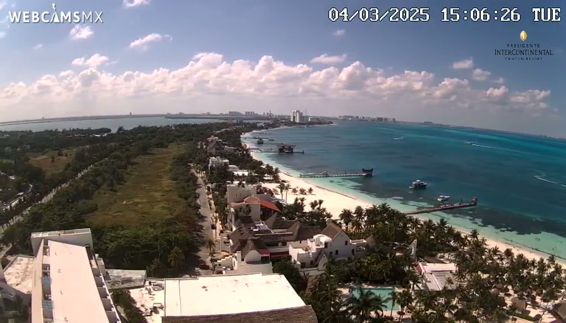 An aerial view of a beach town on a partly cloudy day, showing a white sand beach, turquoise water, palm trees, buildings, and a road.
