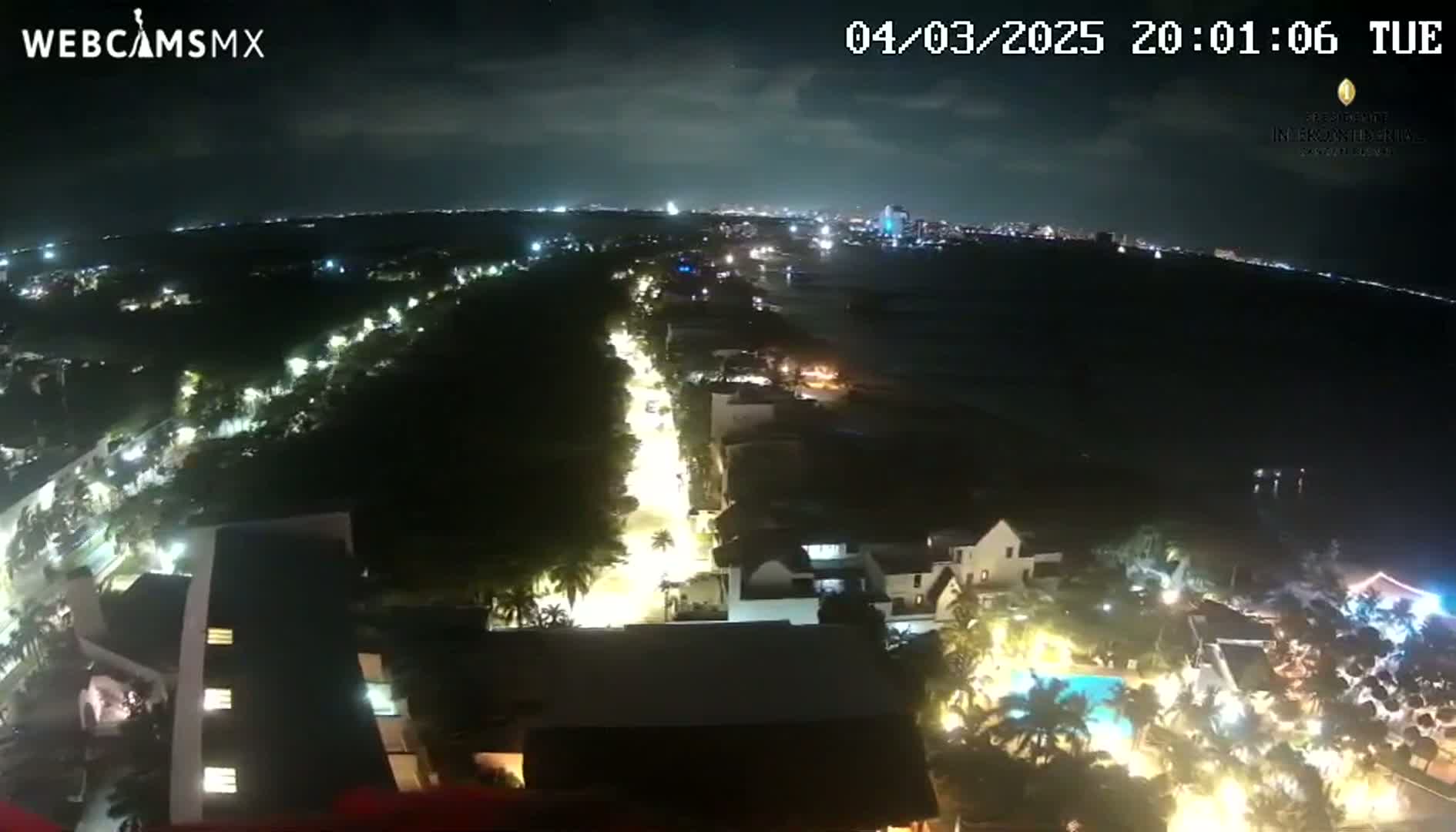 A nighttime, high-angle view shows a brightly lit coastal town with a street lined with lights leading to a dark ocean under a partly cloudy sky.
