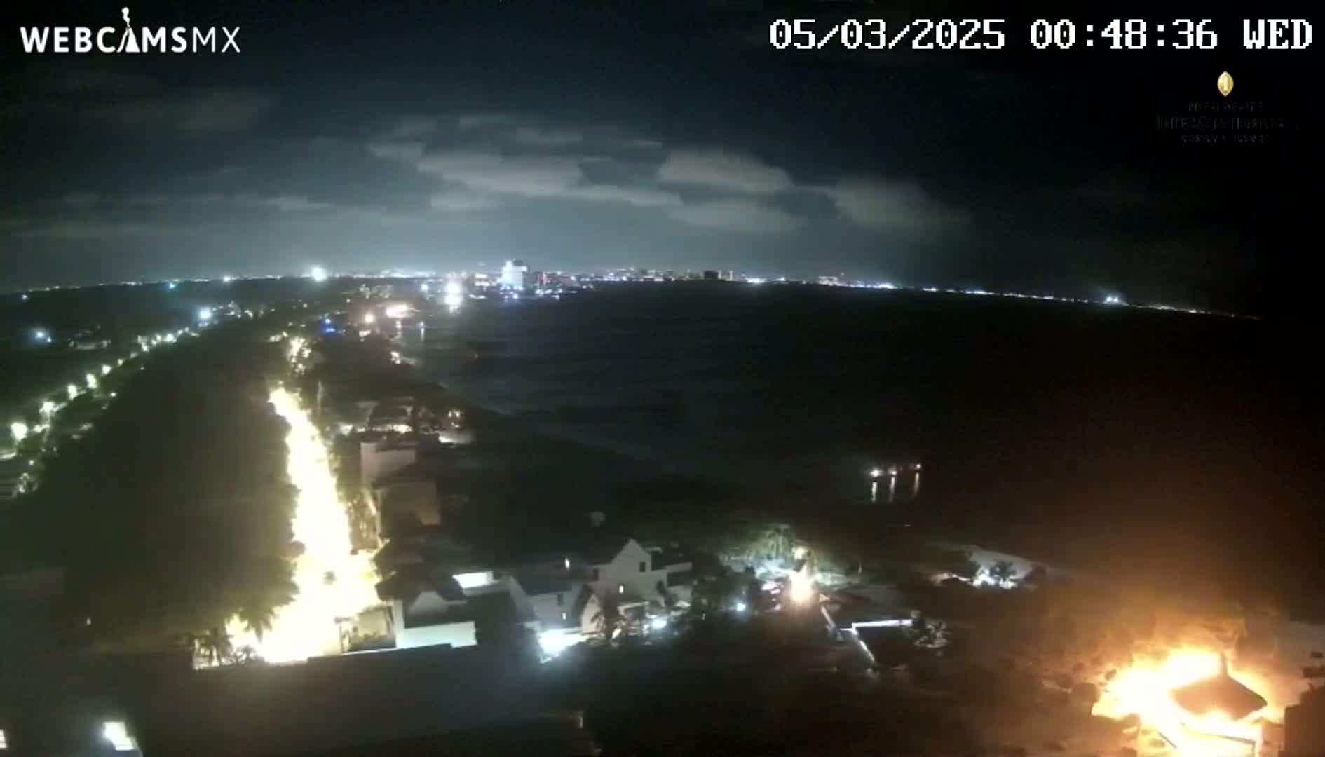 A nighttime aerial view shows a brightly lit beachfront road leading to a city skyline across a dark body of water, with a large fire burning near the bottom right.