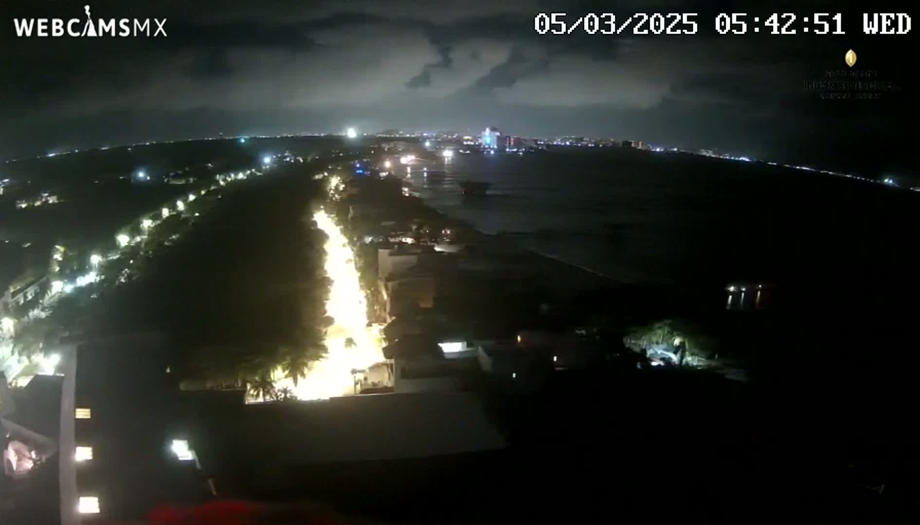 A nighttime, high-angle view shows a brightly lit road next to a dark body of water under a partly cloudy sky.
