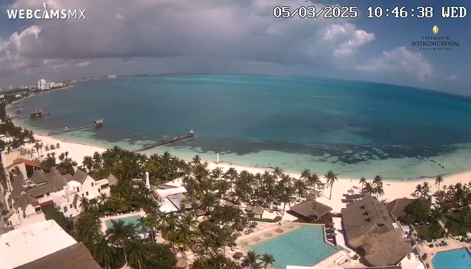 An aerial view shows a tropical beach with turquoise water under partly cloudy skies, featuring a resort with palm trees and swimming pools.