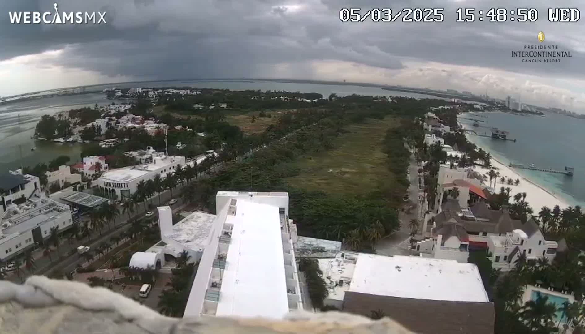 An aerial view shows a coastal town under a cloudy sky, featuring white buildings, lush green vegetation, and a sandy beach bordering a body of water.