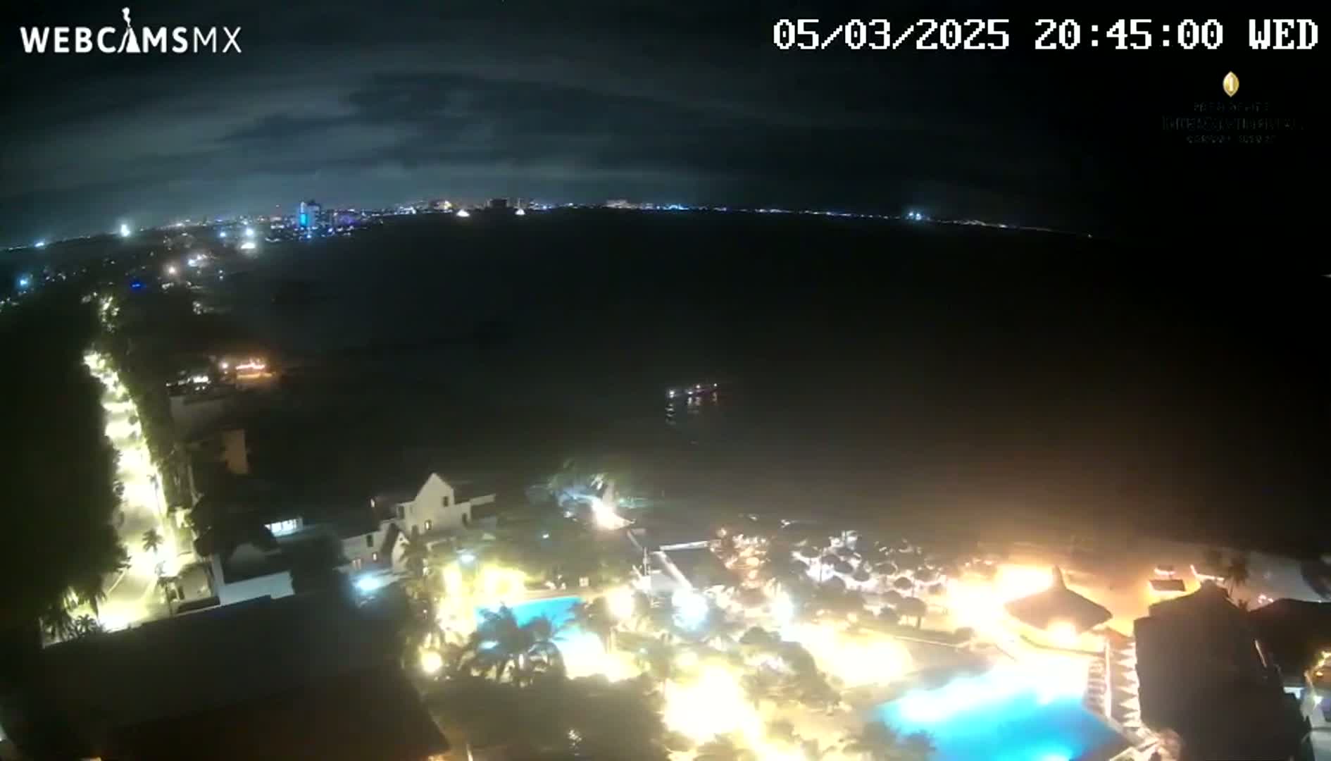 A nighttime aerial view shows a brightly lit resort complex near a body of water with a city skyline visible in the distance under a partly cloudy sky.