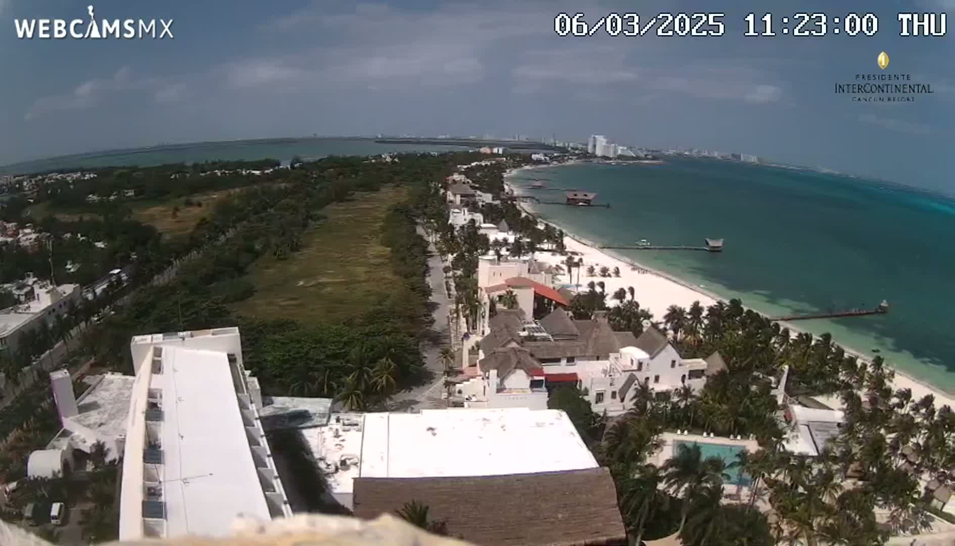An aerial view shows a beach with turquoise water under a partly cloudy sky, featuring buildings and palm trees along the shoreline.
