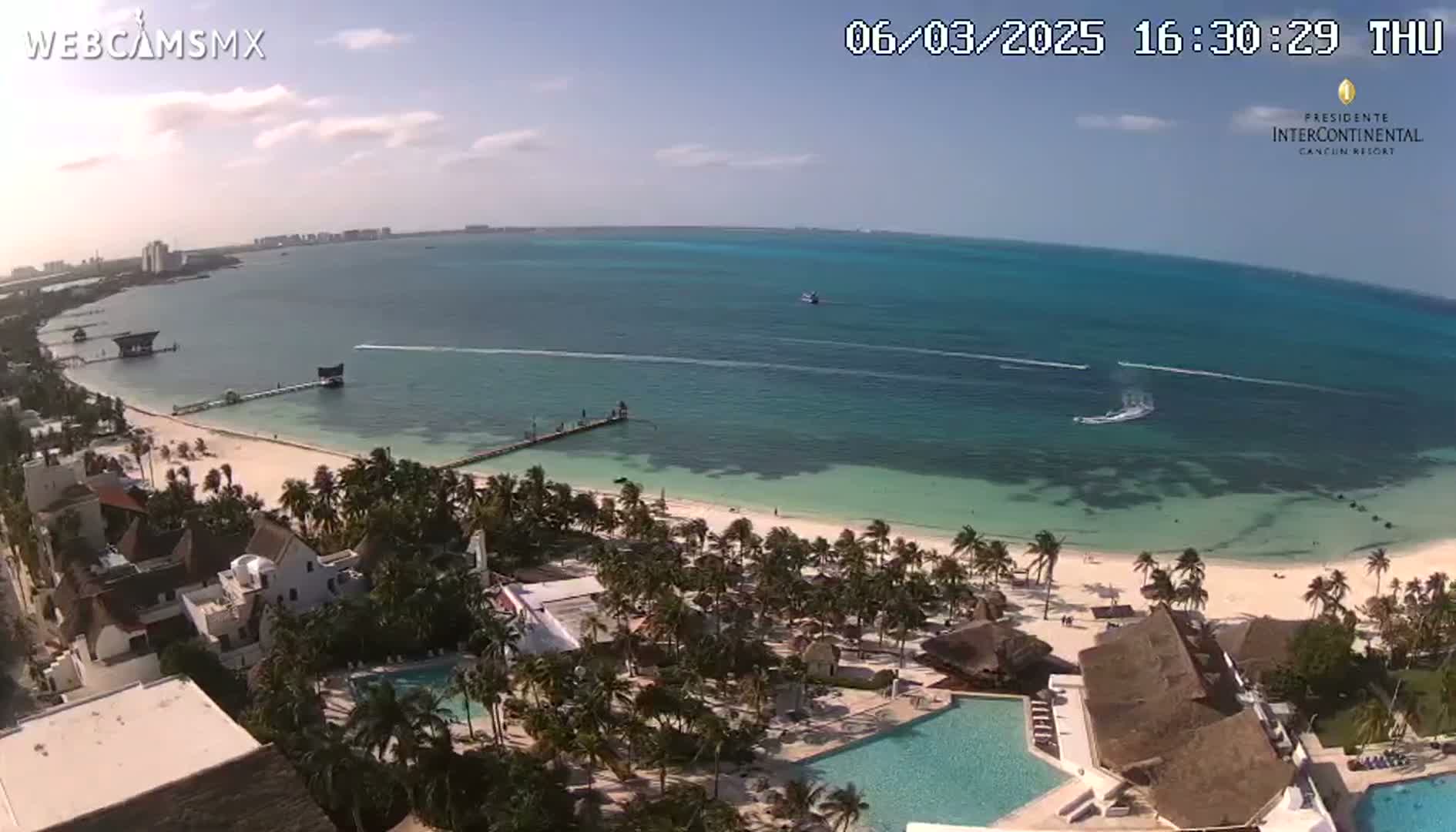 An aerial view shows a tropical beach with turquoise water, palm trees, and a resort with swimming pools under sunny skies.
