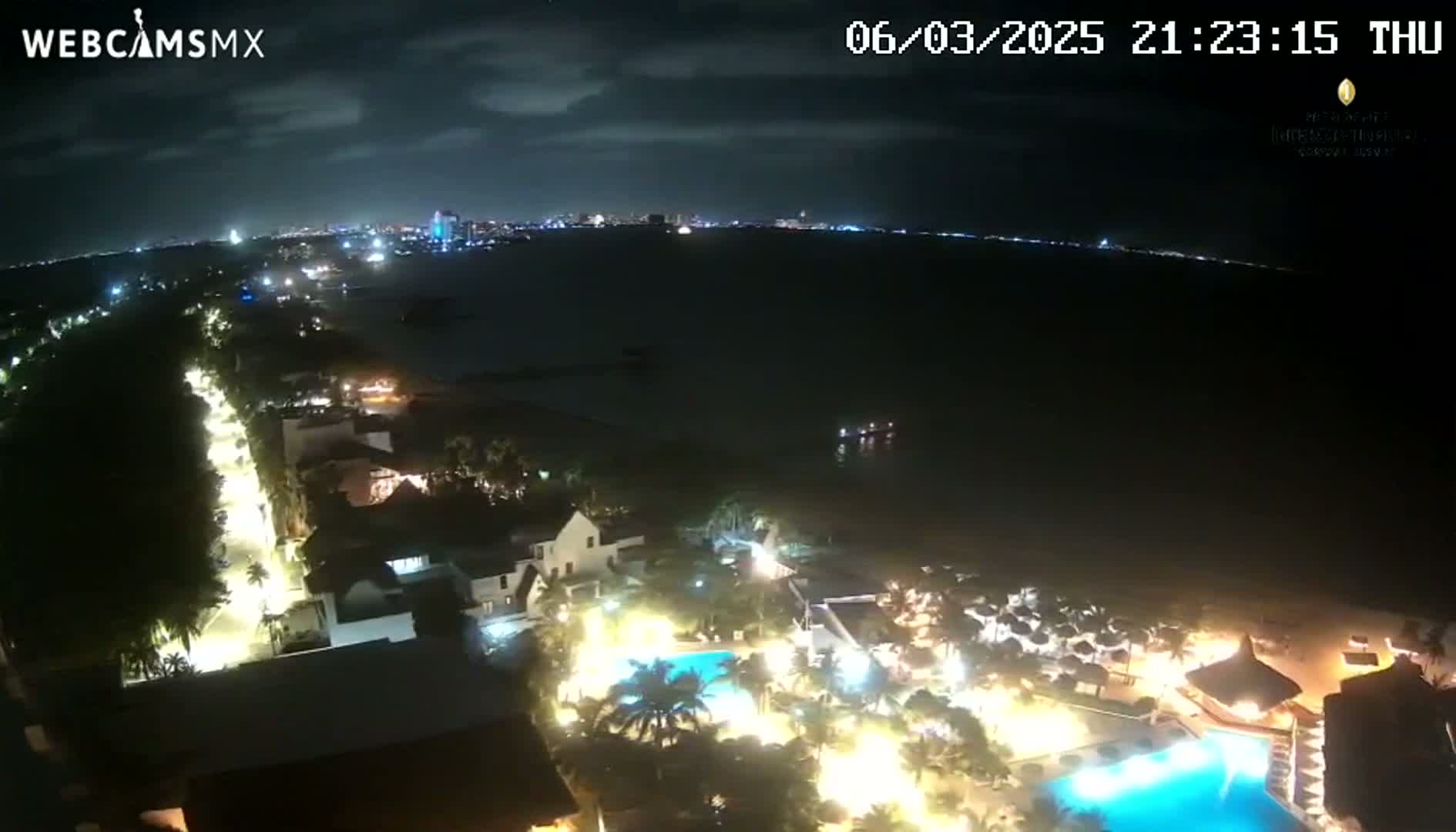 A nighttime aerial view shows a resort complex illuminated with lights, next to a dark body of water with a city skyline visible in the distance under a partly cloudy sky.