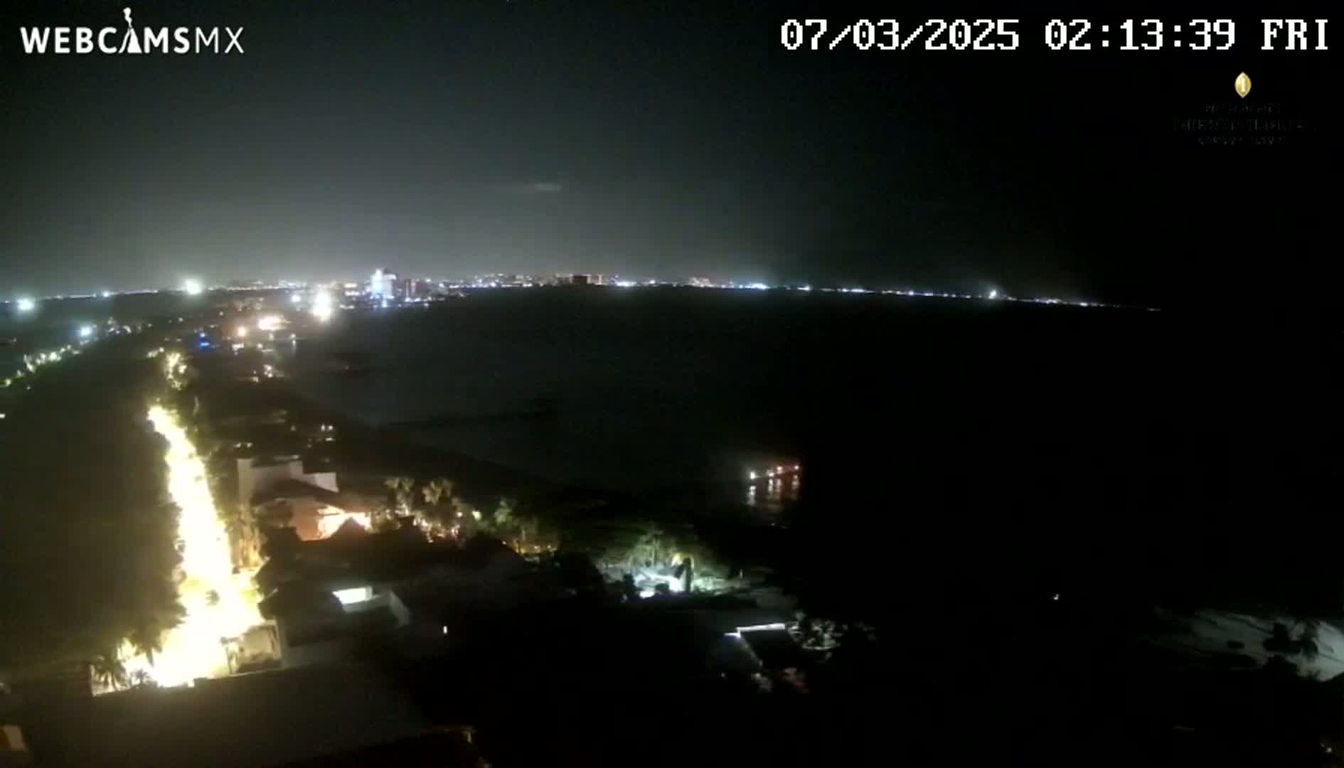 A nighttime coastal scene shows a brightly lit road curving along a dark shoreline, leading towards a distant city skyline under a clear, dark sky.