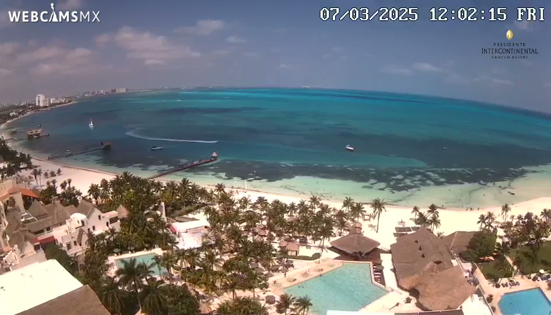 An aerial view shows a resort on a white sand beach next to a calm, turquoise ocean under a mostly sunny sky.