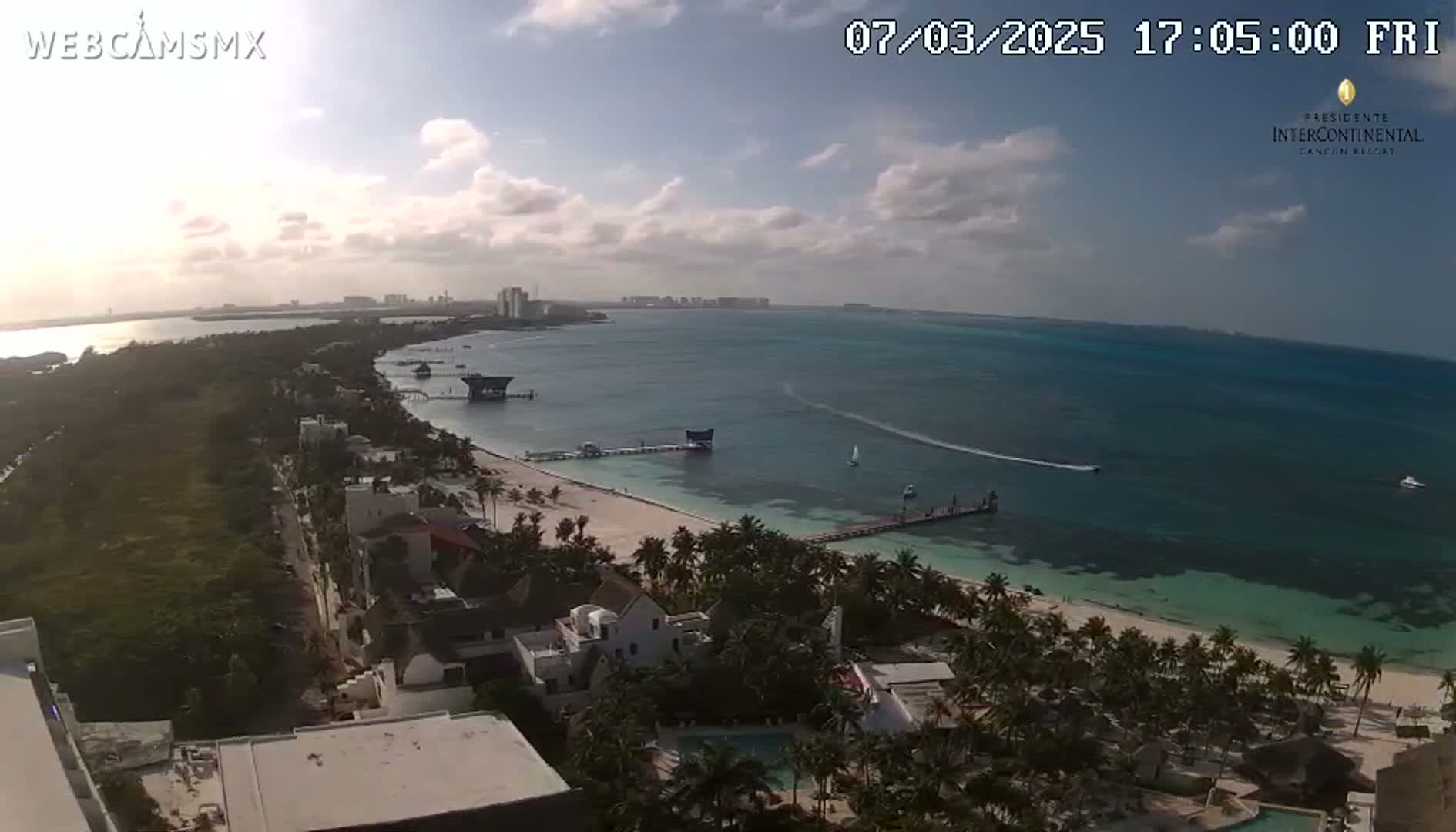 An aerial view shows a partly cloudy day overlooking a beach with turquoise water, palm trees, buildings, and a long pier.