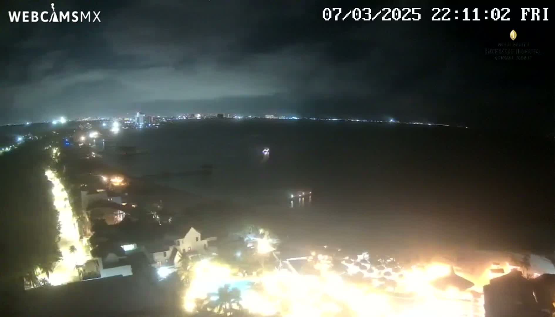 A nighttime, overcast view of a coastal city with brightly lit buildings and a shoreline extending into a dark body of water.